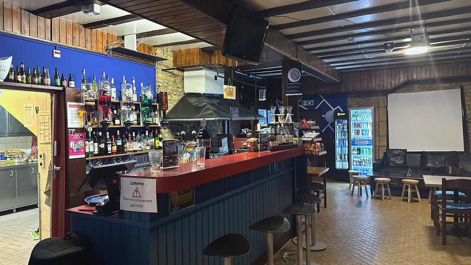 Vue large du Bar Le Forgeron avec son comptoir rouge, bouteilles et tabourets
