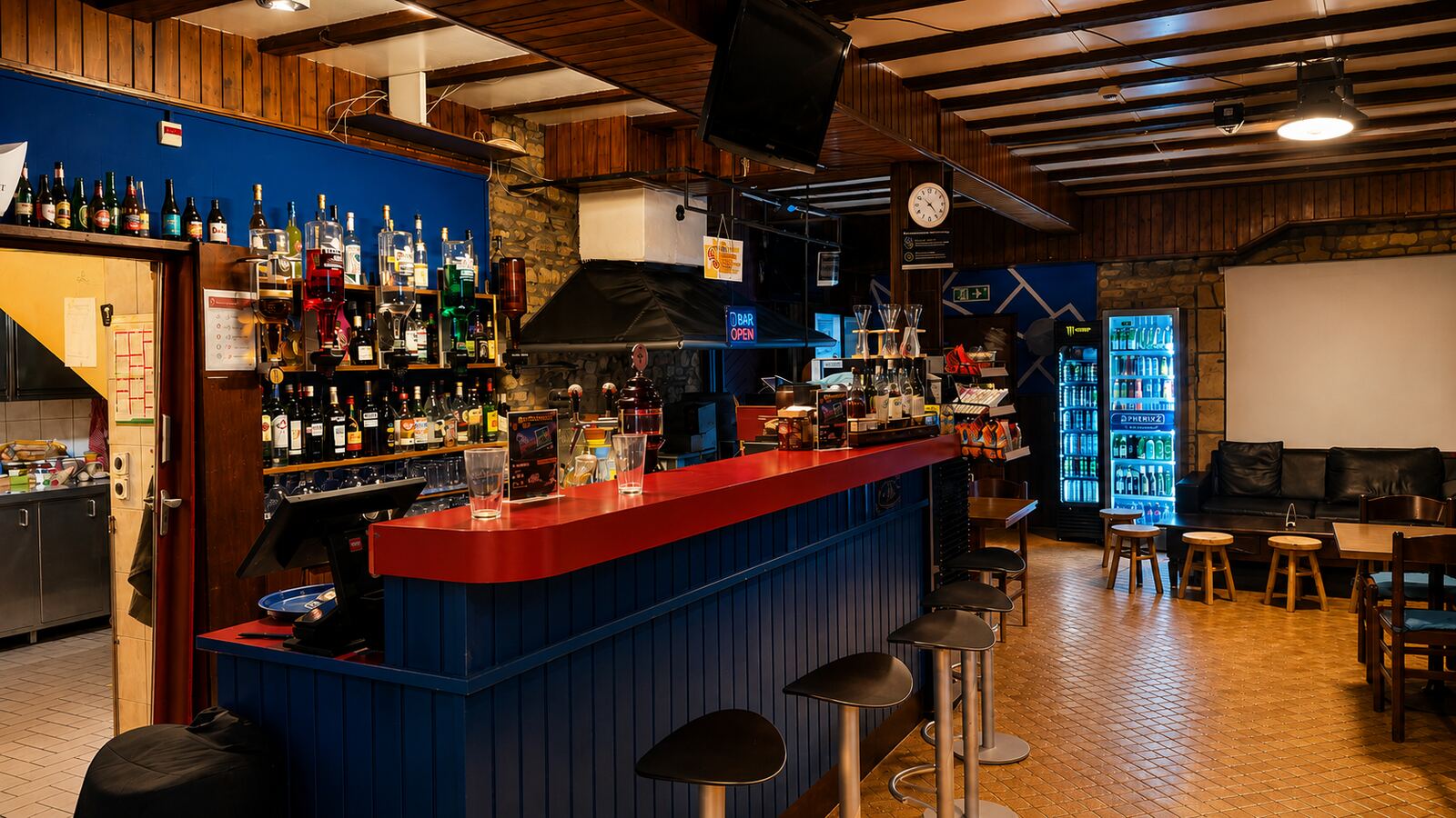 La grande salle : comptoir rouge, frigo à boissons, canapés et écran de projection